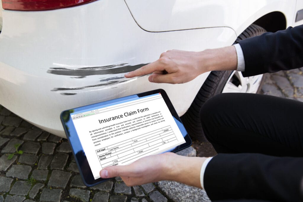 A Person's Hand Examining Damaged Car While Filling Insurance Claim Form On Digital Tablet