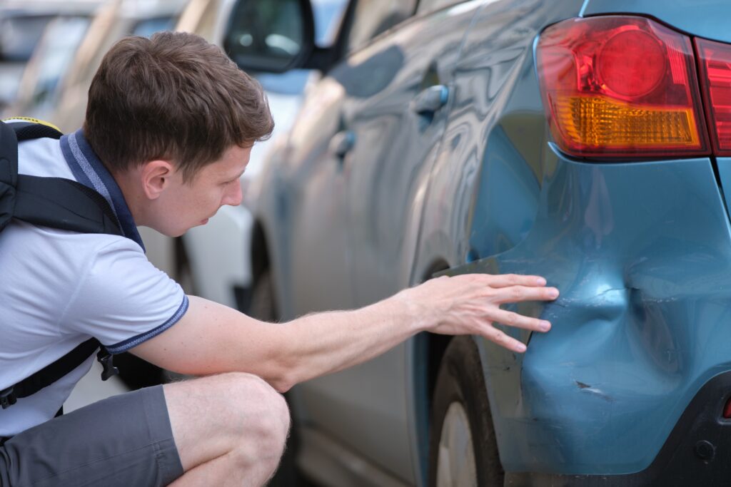 A man examines a large dent and scratches on the rear side of a blue car, likely caused by a collision. Ask ChatGPT