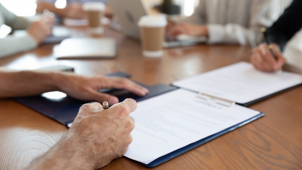 Close-up of people sitting at a table reviewing and signing legal documents with coffee cups and laptops in the background.